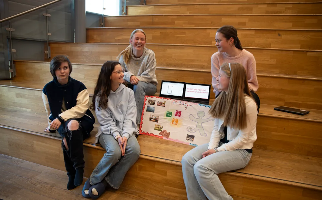 Elevene bak ideen Go social, fotografert sittende i trappa i fellesområdet på skolen med plakaten og presentasjonen av idéen de hadde utviklet i Innovasjonscampen.